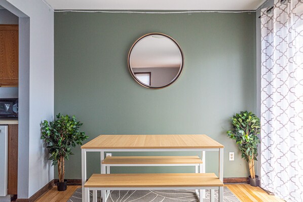 Una habitación con una mesa de madera y un banco, un espejo redondo en la pared y dos plantas en macetas.
