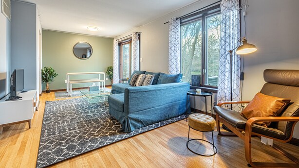 Una sala de estar moderna con un sofá azul, un sillón de cuero marrón, un espejo redondo y un amplio ventanal con cortinas.