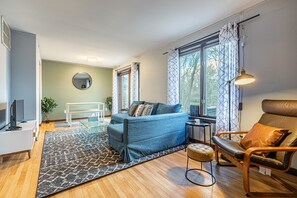 Una sala de estar moderna con un sofá azul, un sillón de cuero marrón, un espejo redondo y un amplio ventanal con cortinas.
