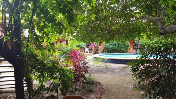 An outdoor patio with a wooden table and wicker chairs, surrounded by lush greenery and a swimming pool. - House located 200 m from the beach, leisure area, restaurants, bars and shops (Mata de São João)