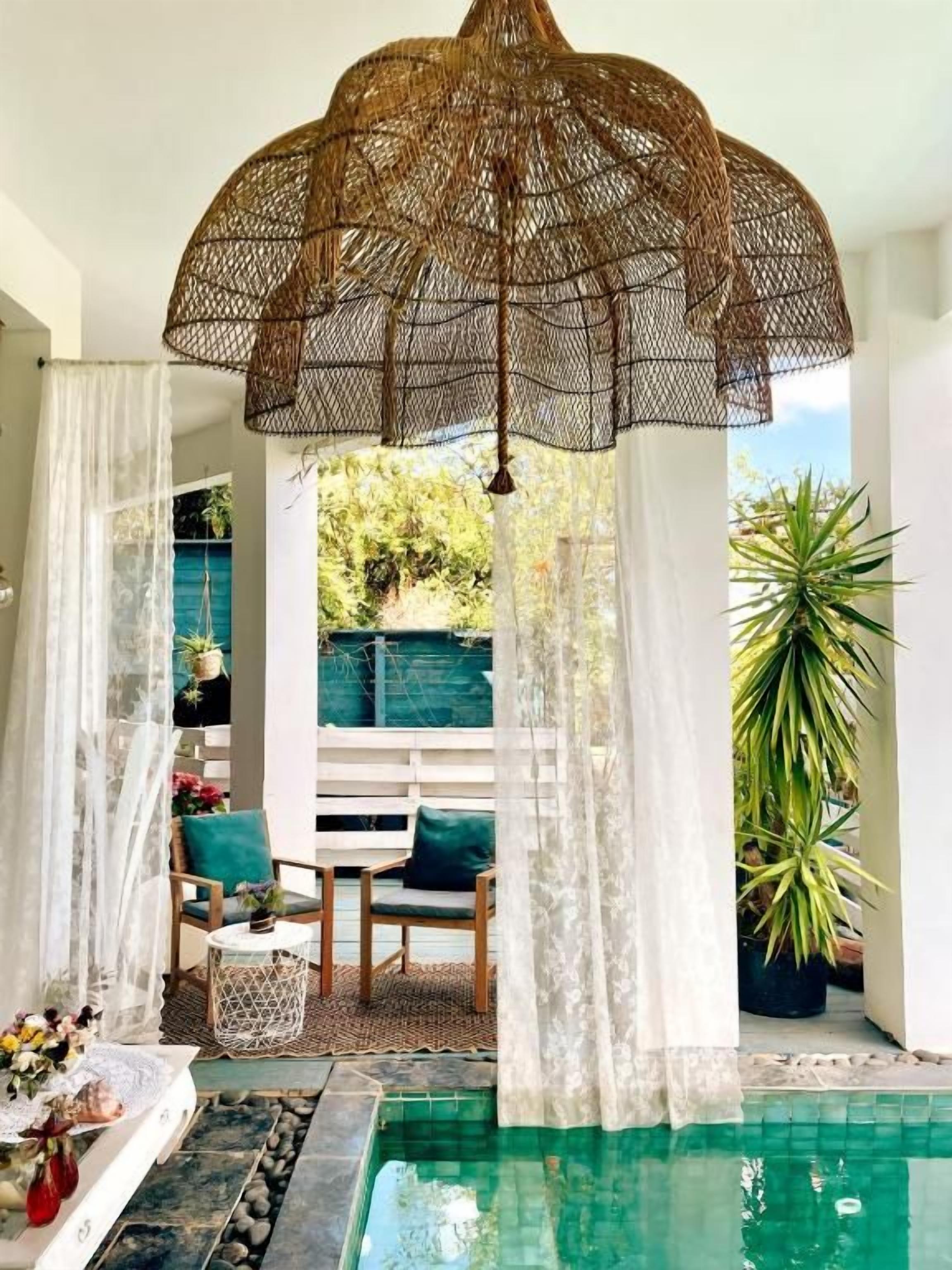 Un espace piscine agrémenté d’un parasol suspendu en rotin, de rideaux blancs et d’un coin salon avec une table et des chaises.