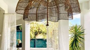 Un espace piscine agrémenté d’un parasol suspendu en rotin, de rideaux blancs et d’un coin salon avec une table et des chaises.