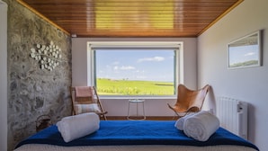 Une chambre avec un mur en pierre, une vue sur un champ verdoyant, un plafond en bois, un lit avec une literie bleue, une chaise et un radiateur.