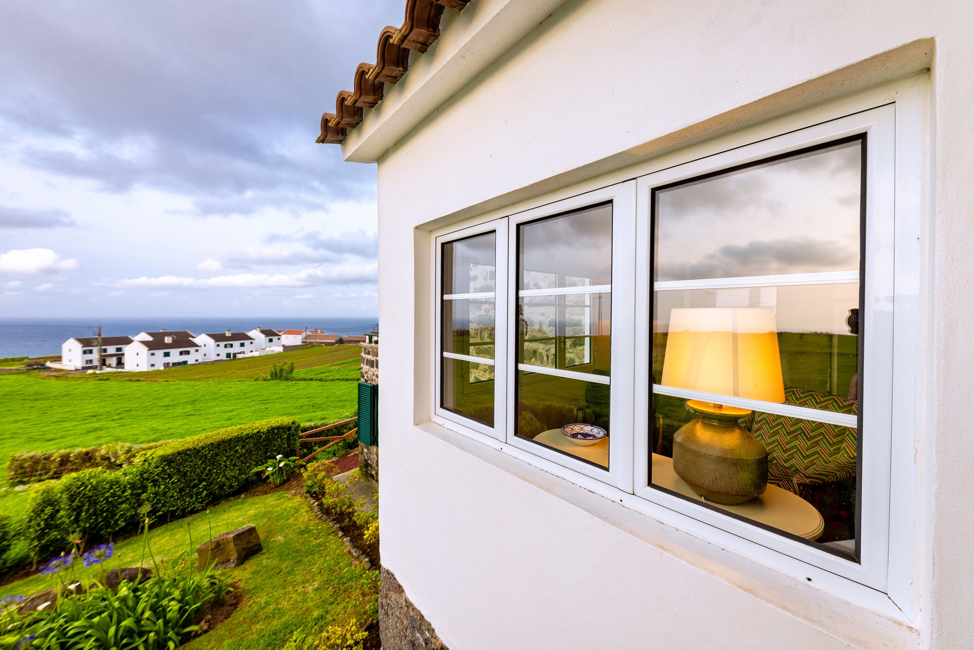 A white building with a window overlooking a green field and a distant view of white houses.