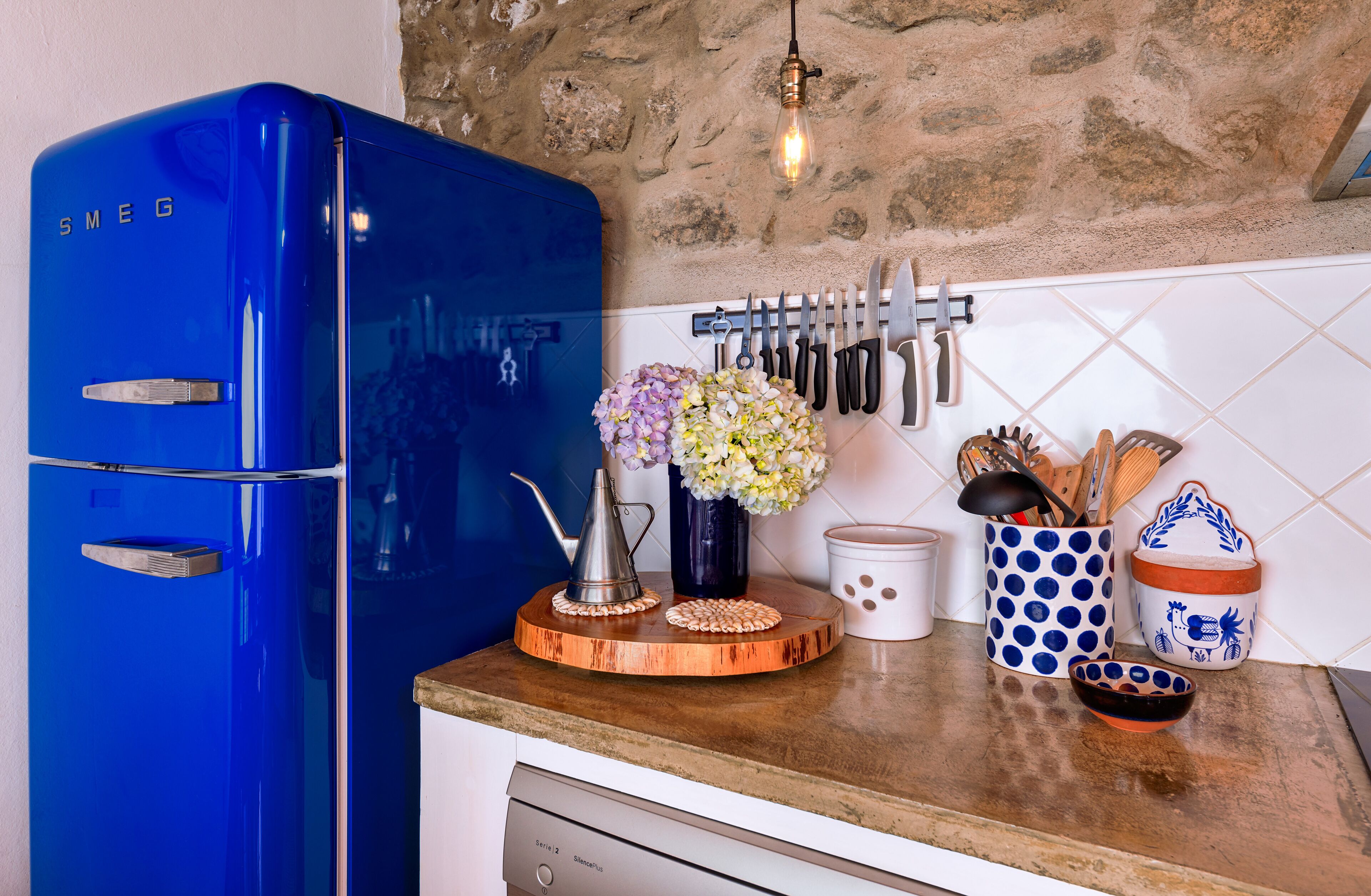 A kitchen with a blue refrigerator, a wooden cutting board, a vase with flowers, and various kitchen utensils.