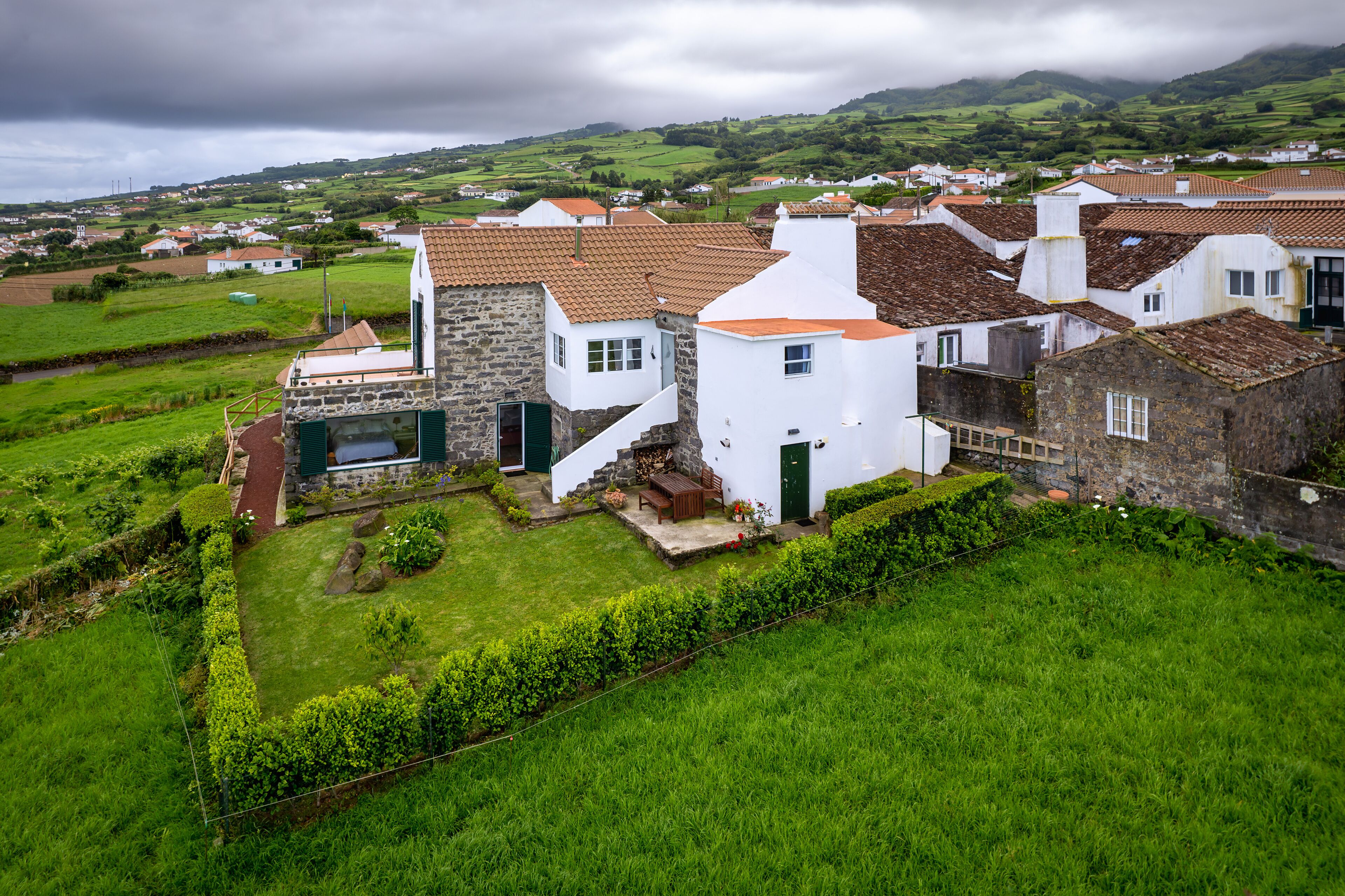 A traditional stone house with a sloped roof, a small garden, and a view of a green valley.