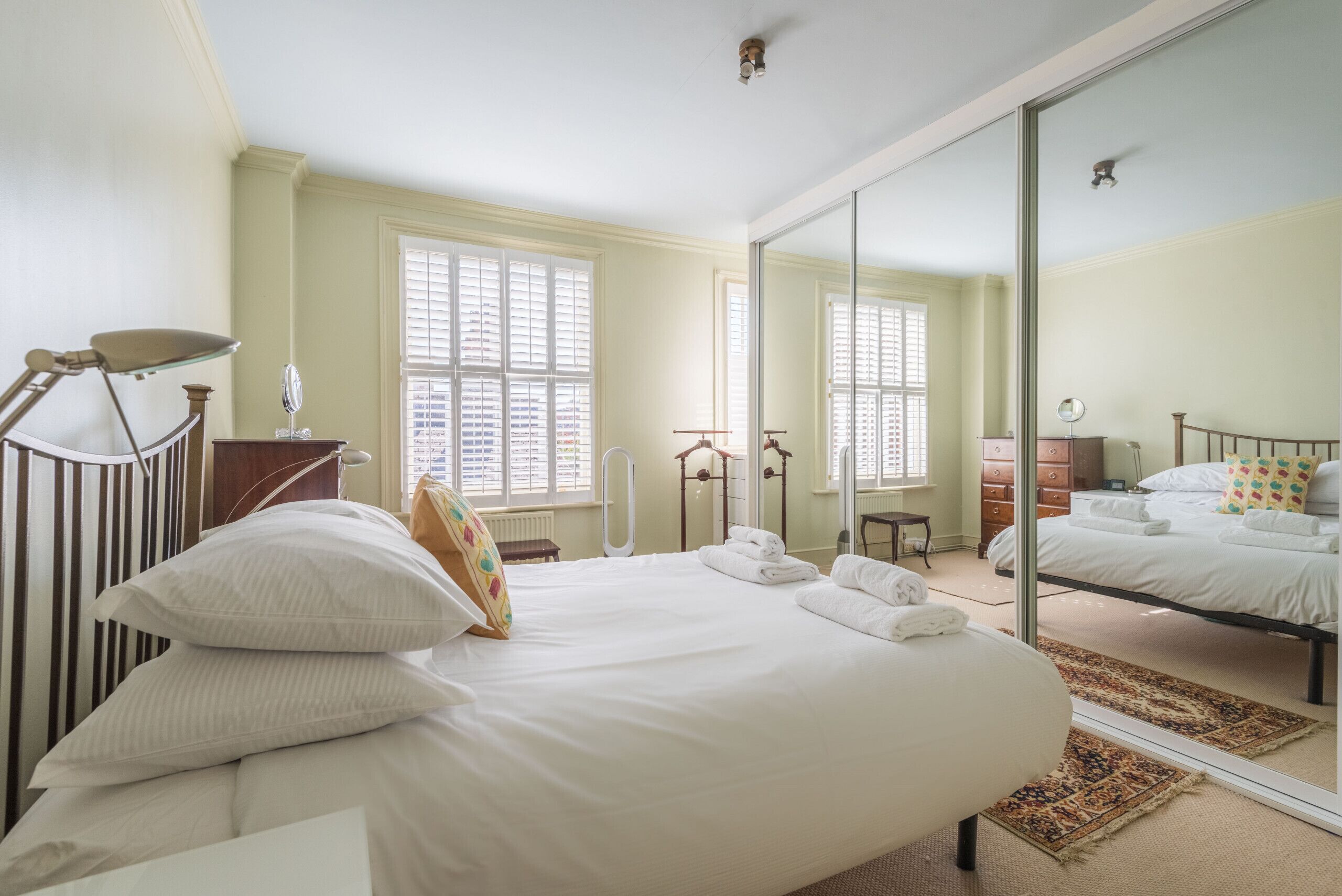 A bedroom with a large bed, a desk, and a chair. There are two windows with blinds, a mirror, and a dresser.