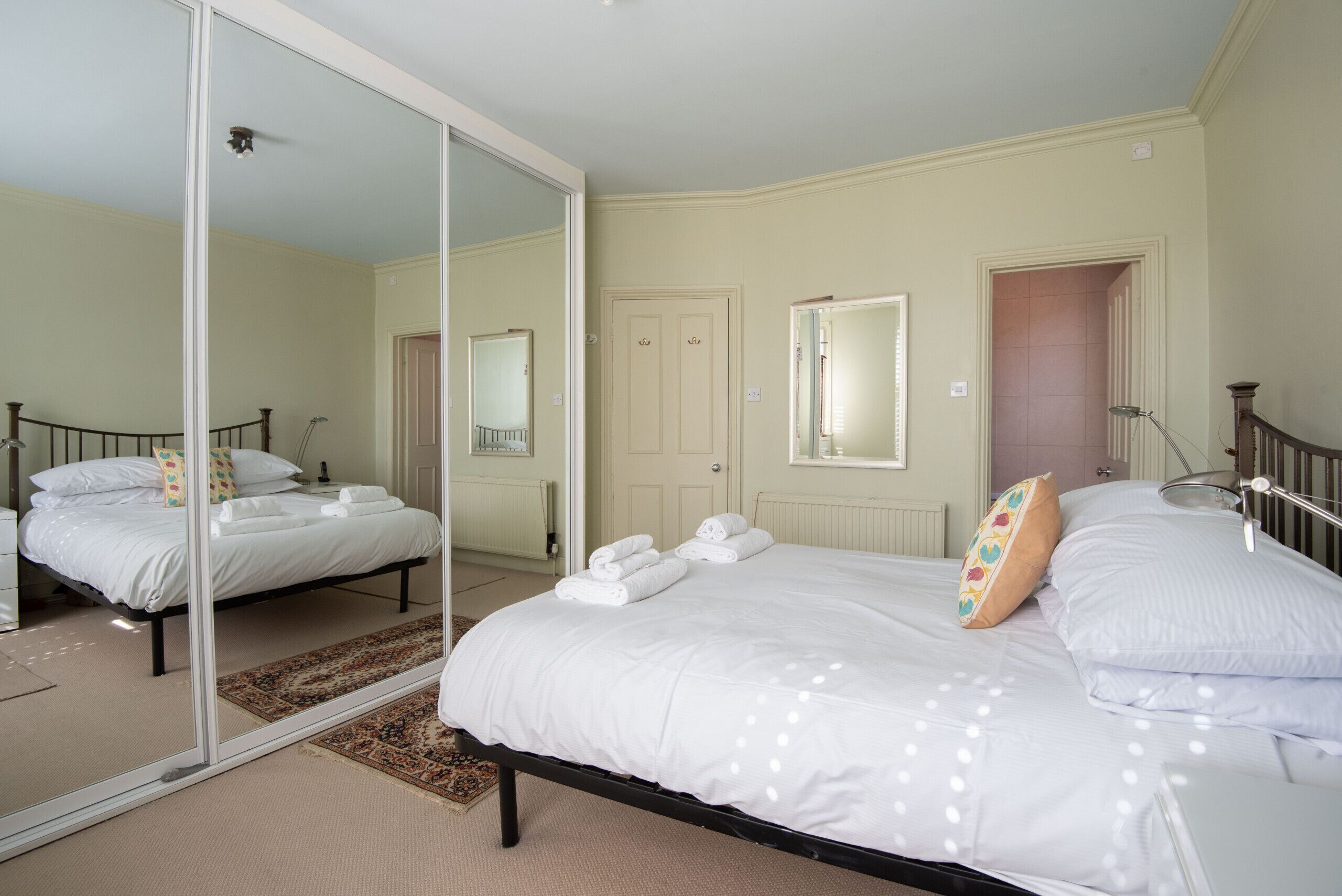 Une chambre avec un grand miroir, un lit recouvert de draps blancs, une table de chevet et une porte.