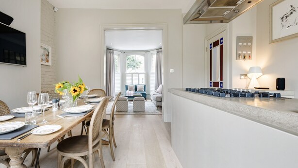A modern kitchen with a central island, a dining area with a wooden table and chairs, and a view into the living room. - A Home on Northcote (London)
