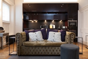 A cozy living room with a tufted sofa, a coffee table, and a dark wooden kitchen in the background.