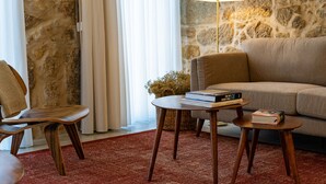 A cozy living room with a stone wall, a sofa, a wooden chair, a small table with books, and a floor lamp. - Portugal Boutique Winery Apartments (Vila Nova de Gaia)