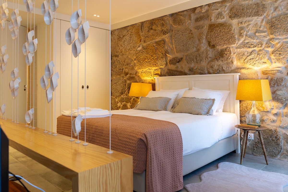 A bedroom with a stone wall, a bed with white linens, two bedside lamps, and a wooden bedside table.