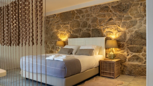 A bedroom with a stone wall, a bed with white linens, two bedside lamps, and a wooden nightstand. - Portugal Boutique Winery Apartments (Vila Nova de Gaia)