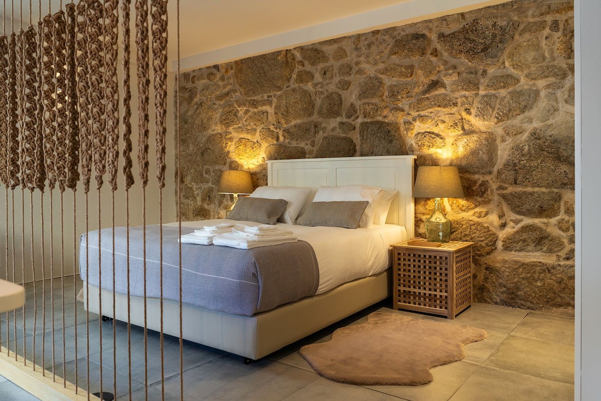 A bedroom with a stone wall, a bed with white linens, two bedside lamps, and a wooden nightstand.