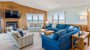 A spacious living room with a large blue sofa, a light blue sofa, and a wicker chair. There is a fireplace, a television mounted on the wall, and large windows overlooking the ocean. - Woodshed: Oceanfront, Gazebo, Rec room, Elevator (Kill Devil Hills)