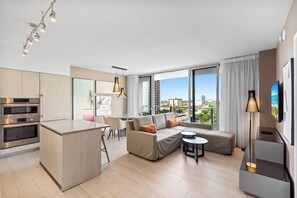 A modern apartment with a kitchen, dining area, and living room. It features a large window with a city view, a sofa with cushions, and a flat-screen TV.