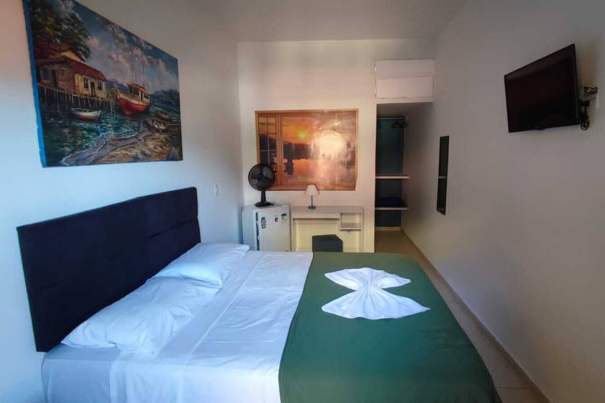 A hotel room with a bed, a green throw blanket, a painting of boats, a TV, and a small kitchenette.