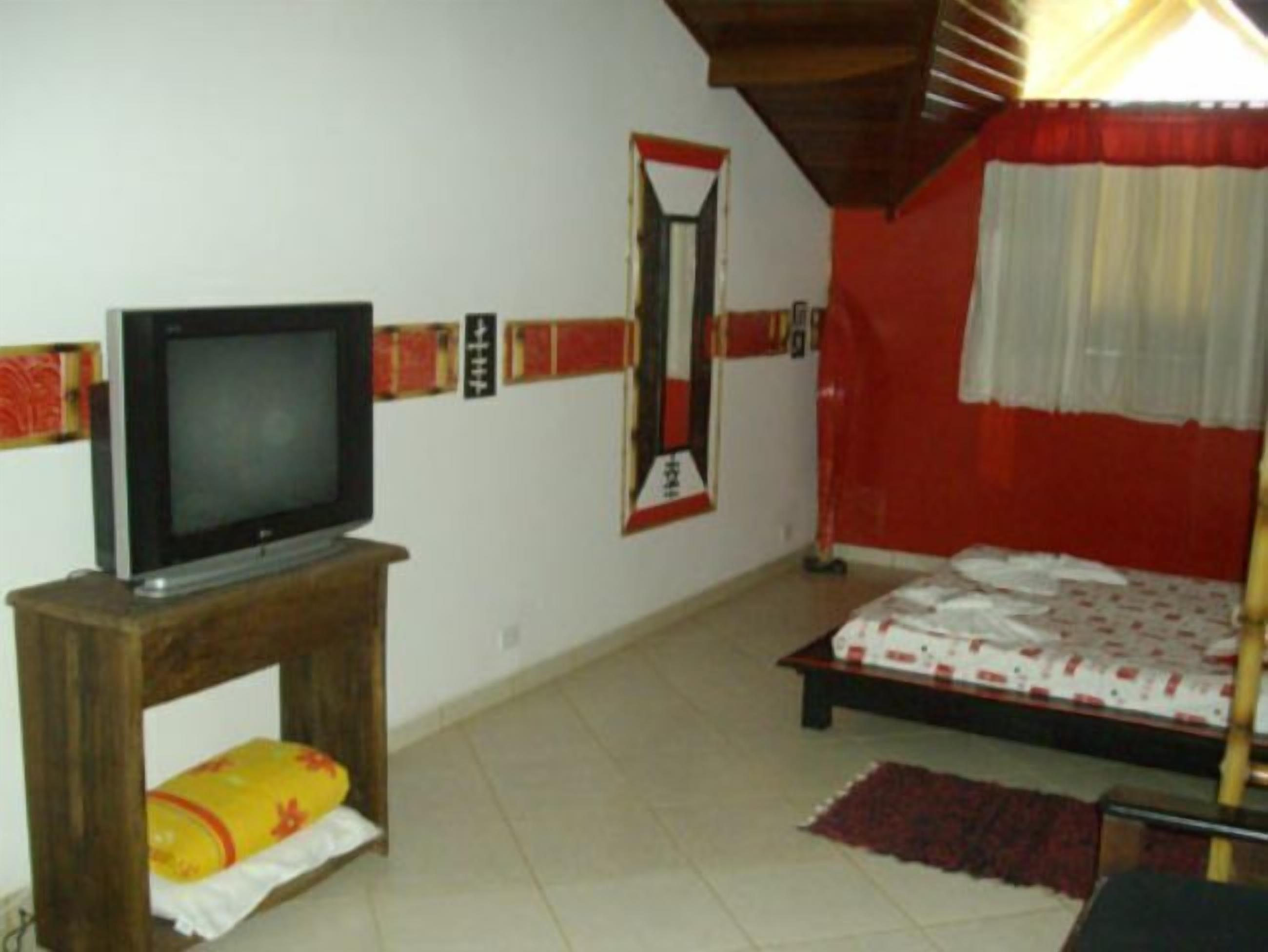 Een kamer met een televisie op een houten standaard, een bed met een bedsprei met een patroon, een spiegel aan de muur, en een rood gordijn.