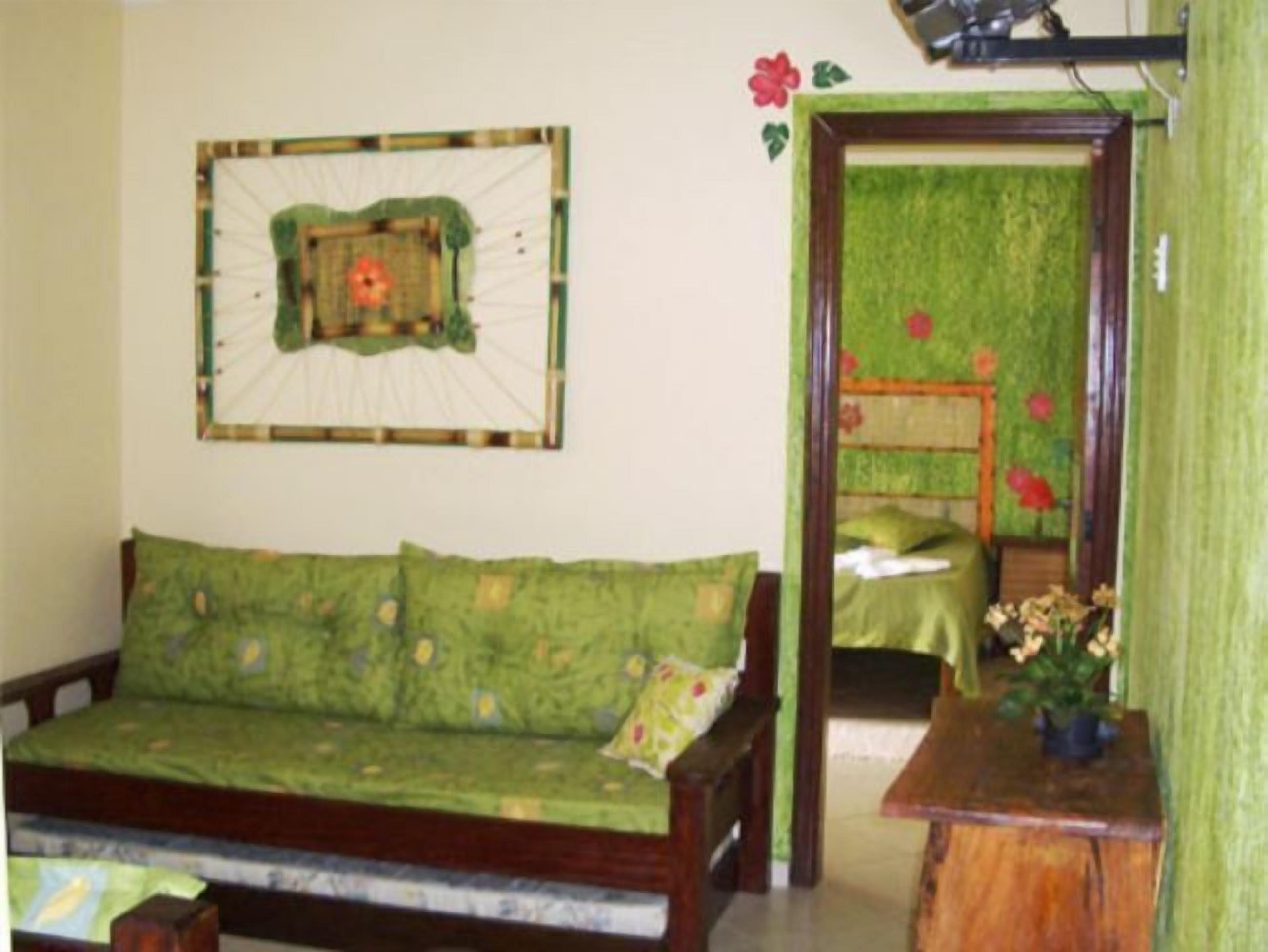 Quarto com sofá verde, uma pequena mesa de madeira com uma planta e uma cama com colcha verde e flores vermelhas.