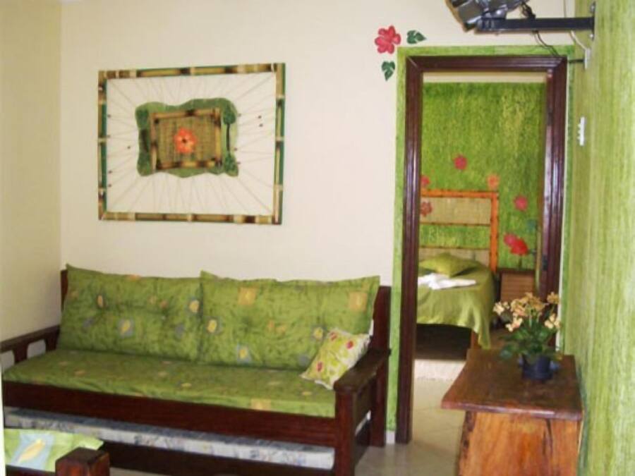 Quarto com sofá verde, uma pequena mesa de madeira com uma planta e uma cama com colcha verde e flores vermelhas.