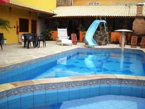 A swimming pool with a blue slide, surrounded by lounge chairs and a tiled deck. - Peruibe Suites Flat (Peruíbe)