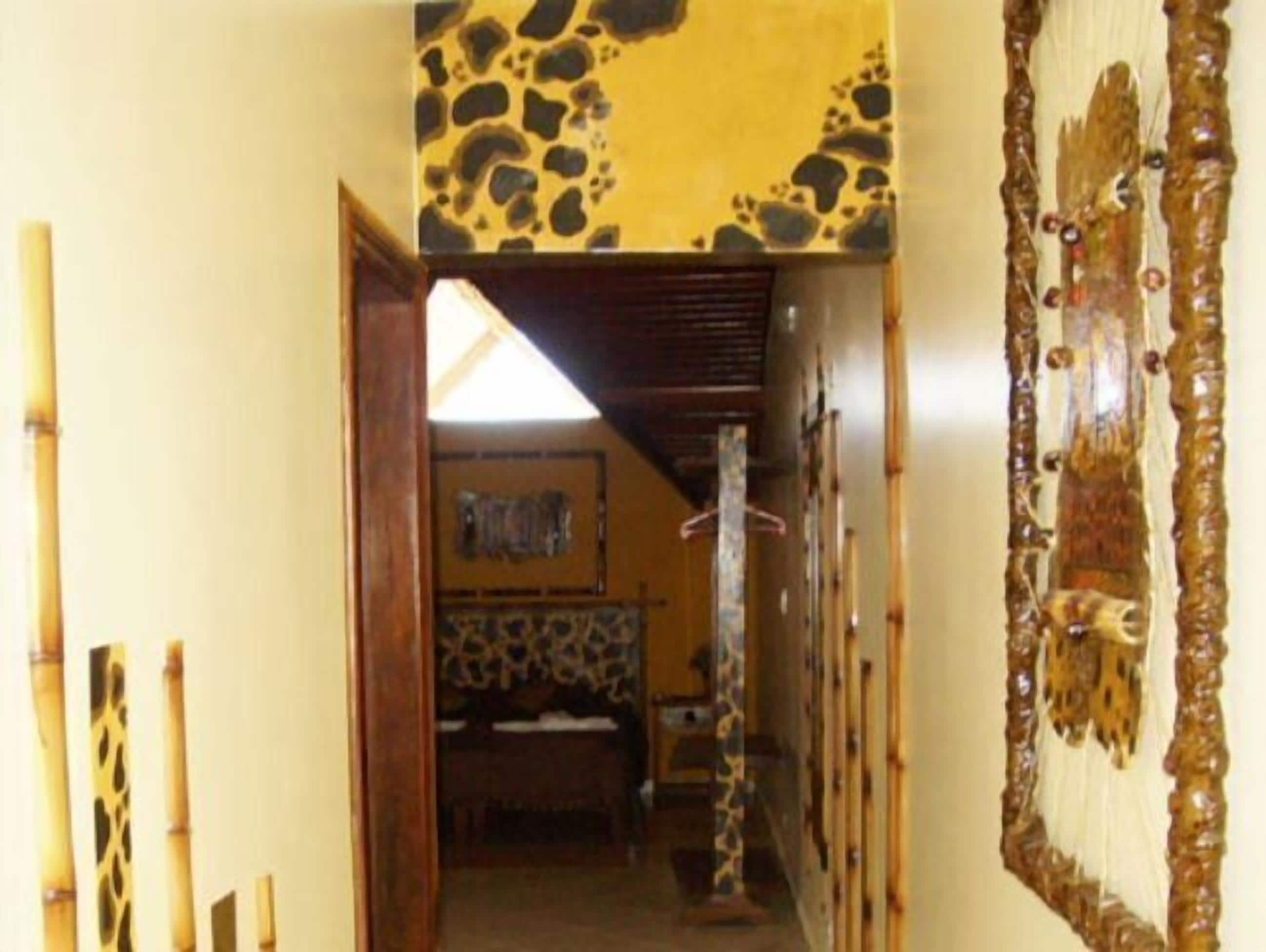 Un pasillo estrecho que conduce a una habitación con una cama y una pared decorada con un diseño. Se ven cañas de bambú a la izquierda y un adorno en la pared a la derecha.