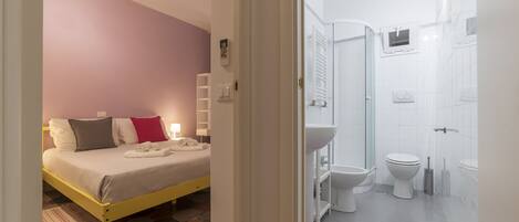 A bedroom with a bed, bedside tables, and a nightstand. The bathroom contains a toilet, sink, and shower.