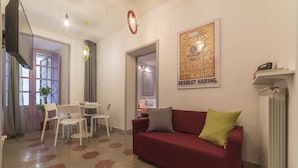 Una sala de estar con un sofá rojo, una mesa de comedor con sillas, un televisor montado en la pared y un radiador.