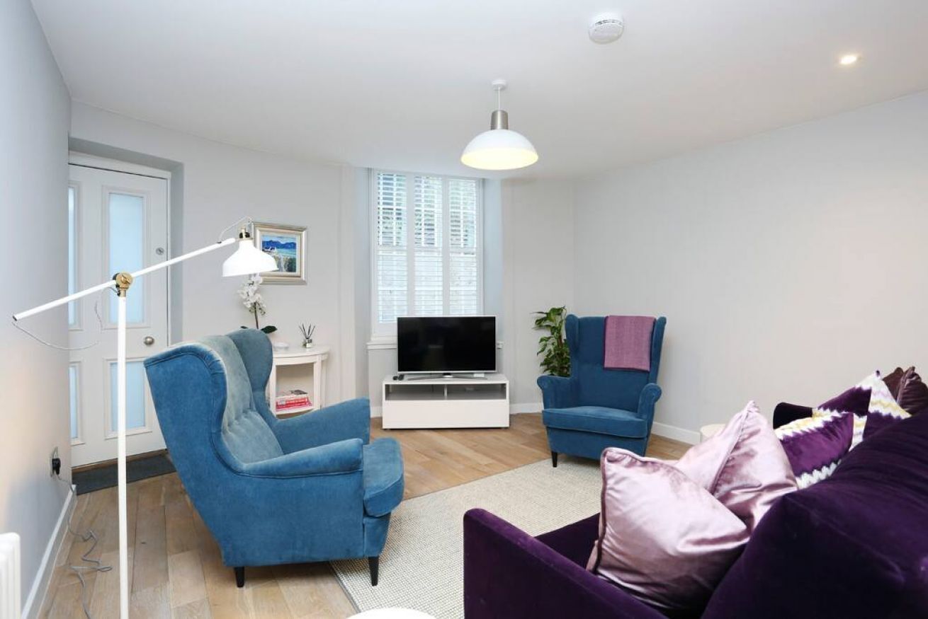 Un salon moderne avec un fauteuil bleu, un canapé violet, un meuble TV blanc et une grande fenêtre avec des stores.
