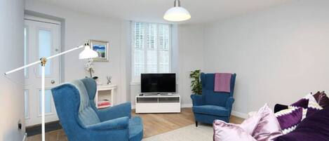 Un salon moderne avec un fauteuil bleu, un canapé violet, un meuble TV blanc et une grande fenêtre avec des stores.
