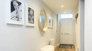 A hallway with framed artwork, a round mirror, a white door, and a small stool with a white cover.