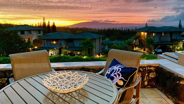 Un patio con una mesa redonda y dos sillas de mimbre, con vista a una zona residencial con casas y un atardecer.