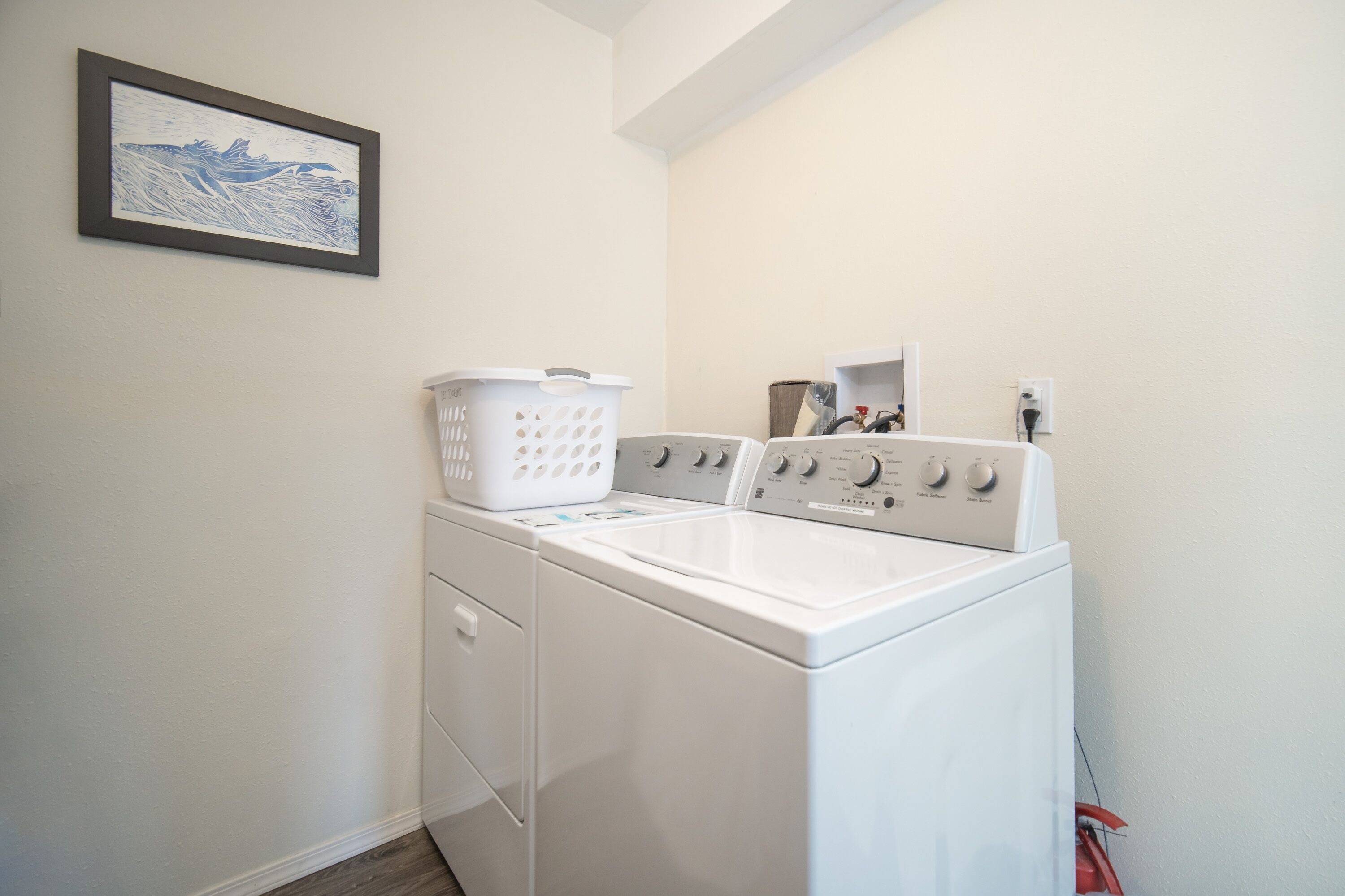 Un espace buanderie équipé d’un lave-linge et d’un sèche-linge à chargement frontal, d’un panier à linge blanc et d’un tableau encadré accroché au mur.