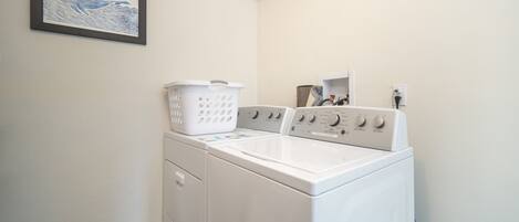 A laundry area with a front-loading washer and dryer, a white laundry basket, and a framed picture on the wall.