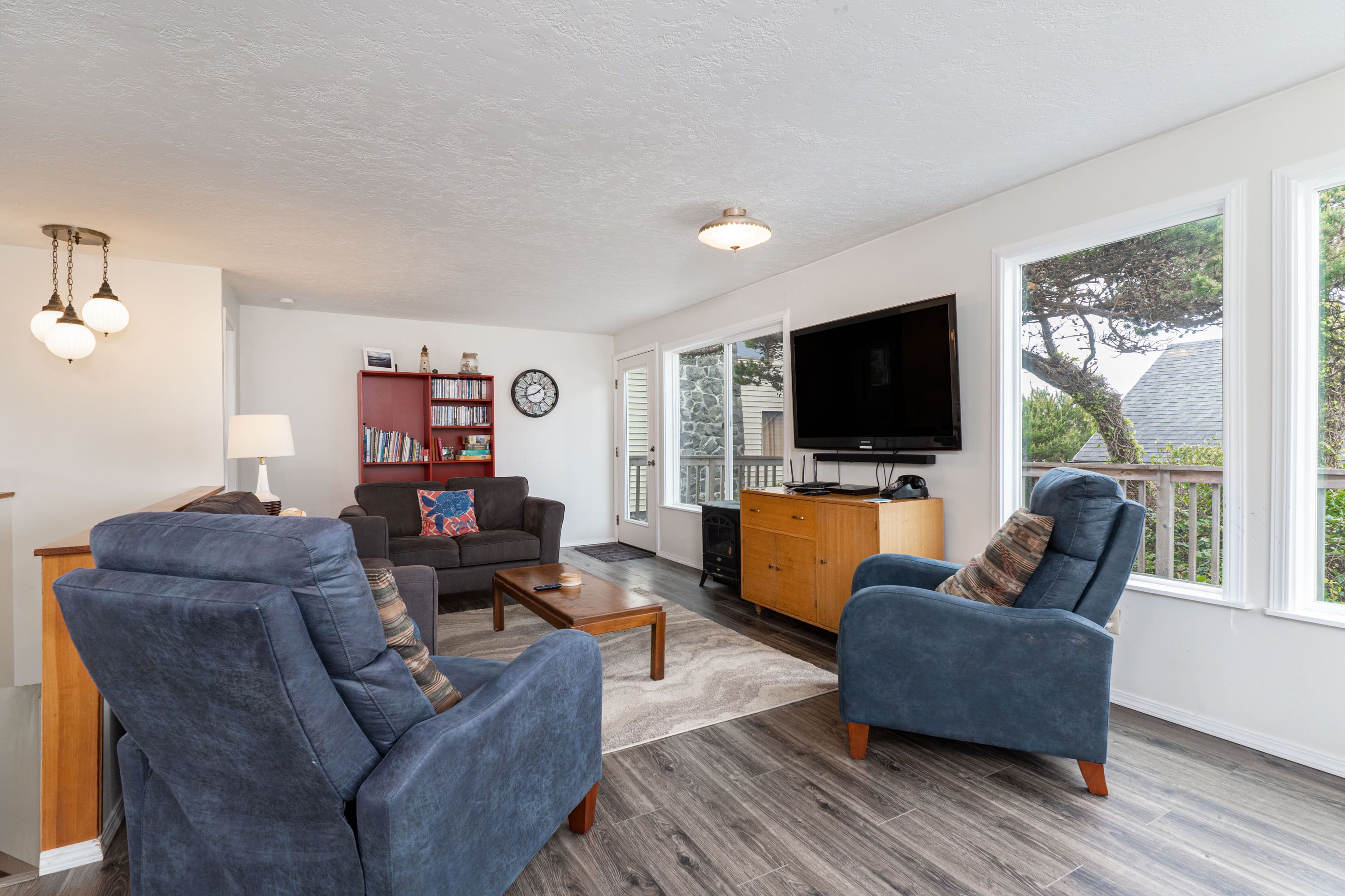 A spacious living room with a large TV, comfortable seating, a bookshelf, and a view through the large windows.