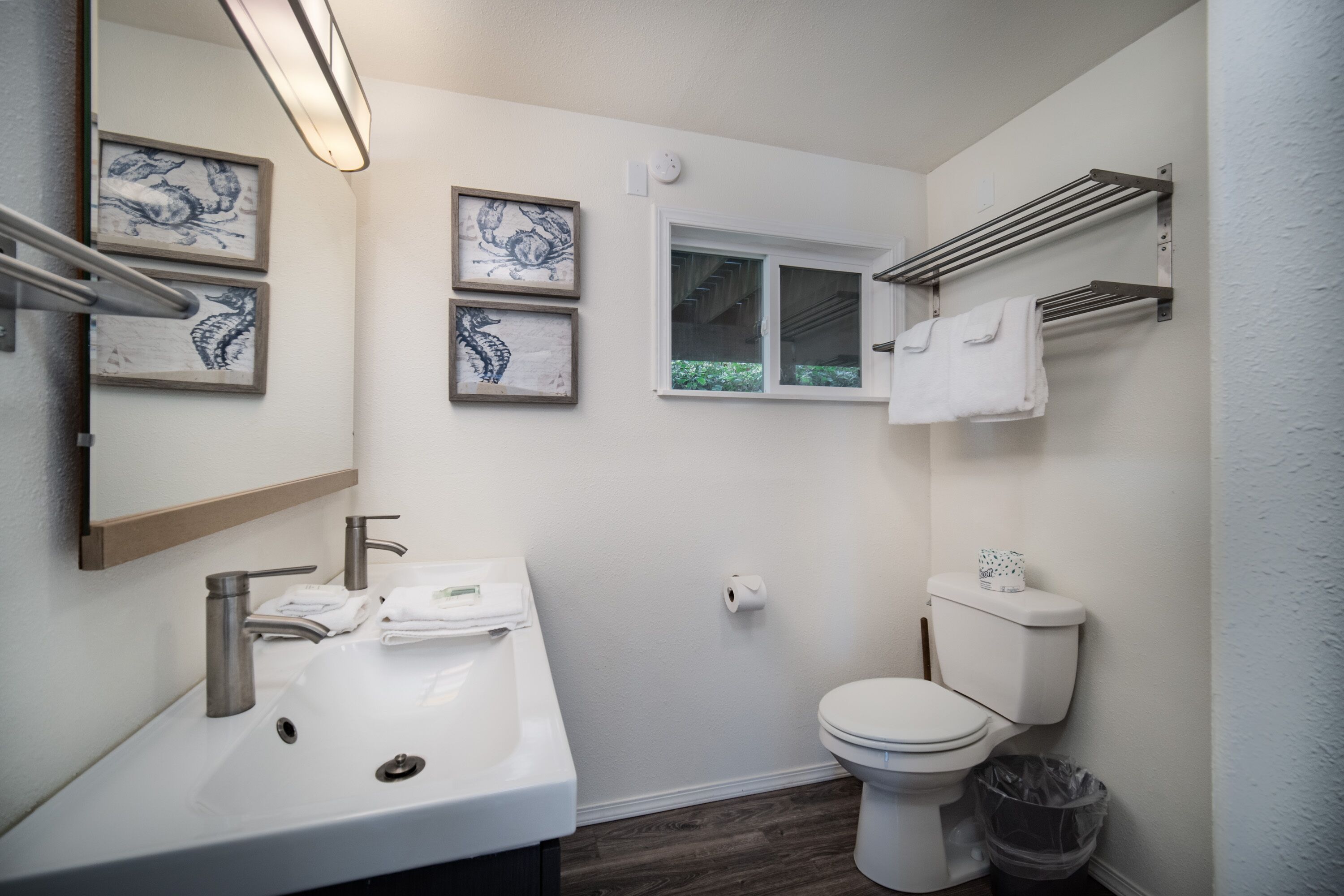 Ein modernes Badezimmer mit einem weißen Waschbecken, zwei Wasserhähnen, einer Toilette und einem Fenster. An den Wänden befinden sich Bilder in Rahmen und ein Handtuchhalter über der Toilette.