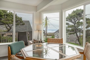 A dining area with a glass table and chairs, large windows with a view of houses and trees, and a lamp on a side table.