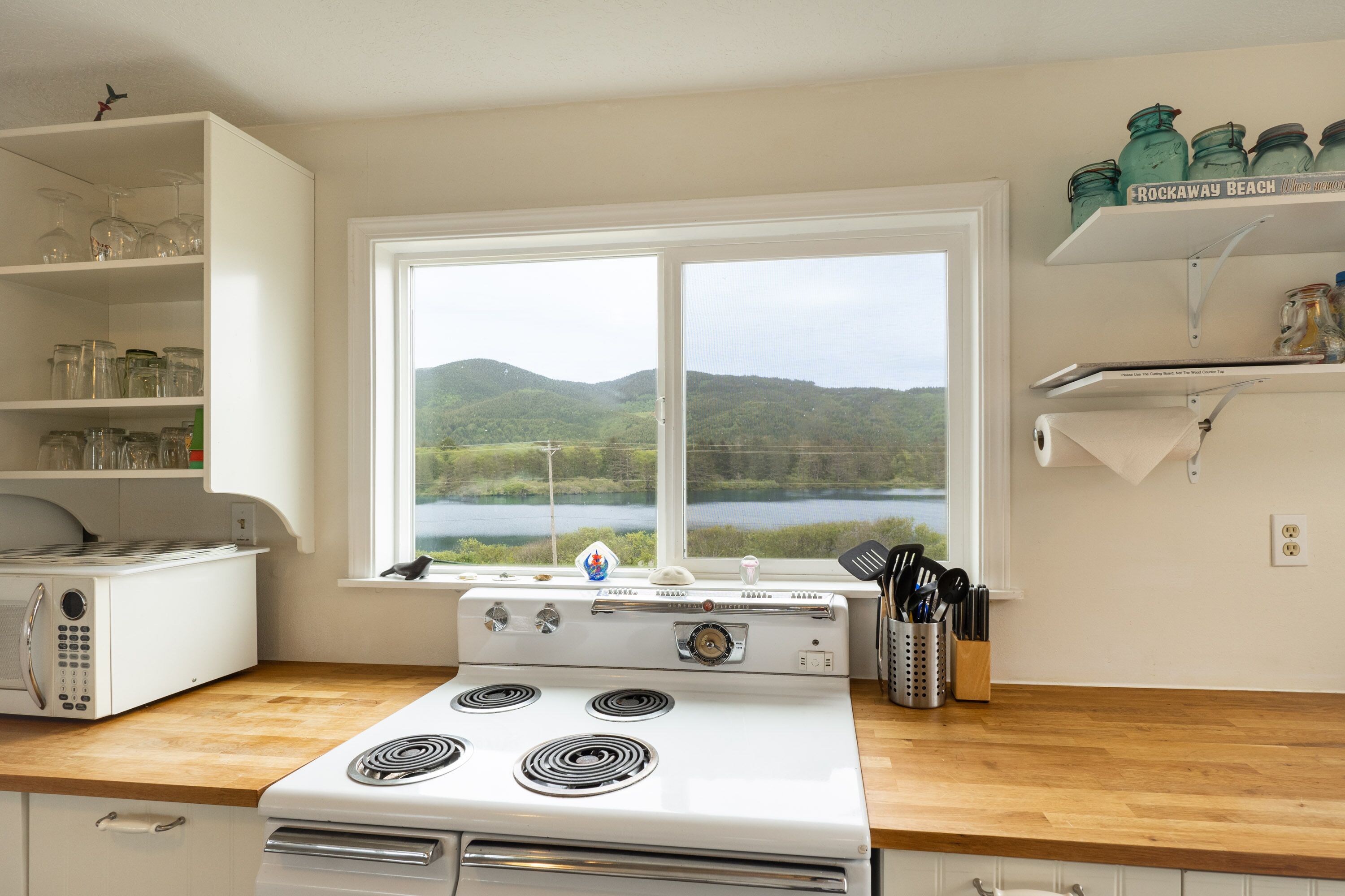 A kitchen with a stove, a microwave, and a window with a view of a lake and hills.