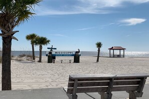 A beach scene with a sign reading 'MISSISSIPPI GULF COAST,' a wooden bench, palm trees, and a gazebo.