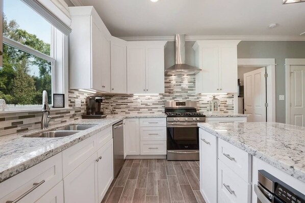 A modern kitchen with white cabinets, a stainless steel range hood, a built-in oven, and a large window providing natural light.