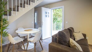 Ein modernes Wohnzimmer mit einer braunen Couch, einem weißen Esstisch mit Stühlen, einer Treppe und einem Balkon mit Blick auf Grünflächen.