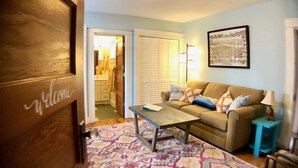 A cozy living room with a sofa, a coffee table, and a patterned rug. There's a framed picture on the wall and a door with a 'Welcome' sign. - ⭐️Tranquil Hidden Gem in the ❤️ of Woodstock⭐️ (Portland)