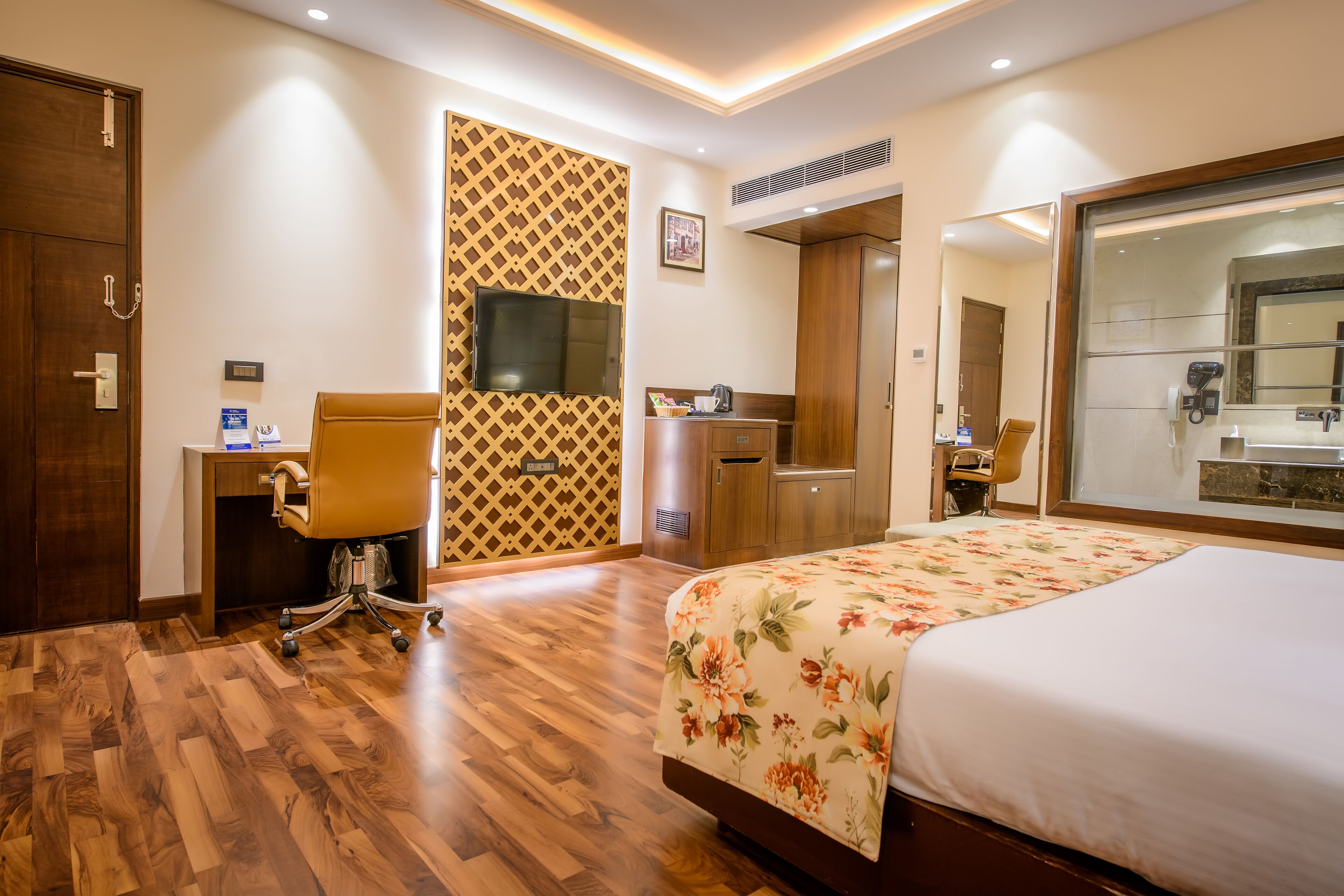 A hotel room with a wooden floor, a bed with a floral bedspread, a desk with a chair, a wardrobe, a television mounted on the wall, and a bathroom with a mirror and sink.