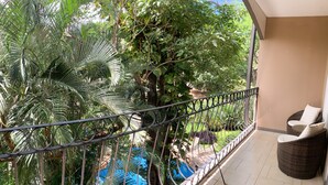 Een balkon met een rieten stoel, een metalen hekwerk en weelderige beplanting op de achtergrond.