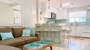 Een moderne woonkamer met een bank, een glazen salontafel en een keukenhoek met witte kasten, een magnetron en een barkruk.