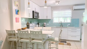 Een moderne keuken met witte kasten, een magnetron, een kookplaat en een spoelbak. Er staan vier stoelen rond een tafel, en een raam met zonwering.
