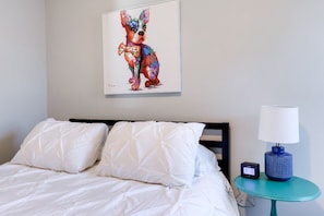 A hotel room with a bed, white bedding, a wall-mounted painting of a dog, a small table with a lamp, and a digital clock.