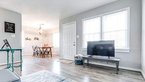 A living room with a TV, a dining area with a table and chairs, and a large window with blinds. - Sunny Cottage At Byrd Pard In Richmond (Richmond)