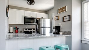 A modern kitchen with white cabinets, a stainless steel refrigerator, a microwave, and a coffee maker. There are three turquoise stools and a wall-mounted sign that reads "Be our guest." - Sunny Cottage At Byrd Pard In Richmond (Richmond)