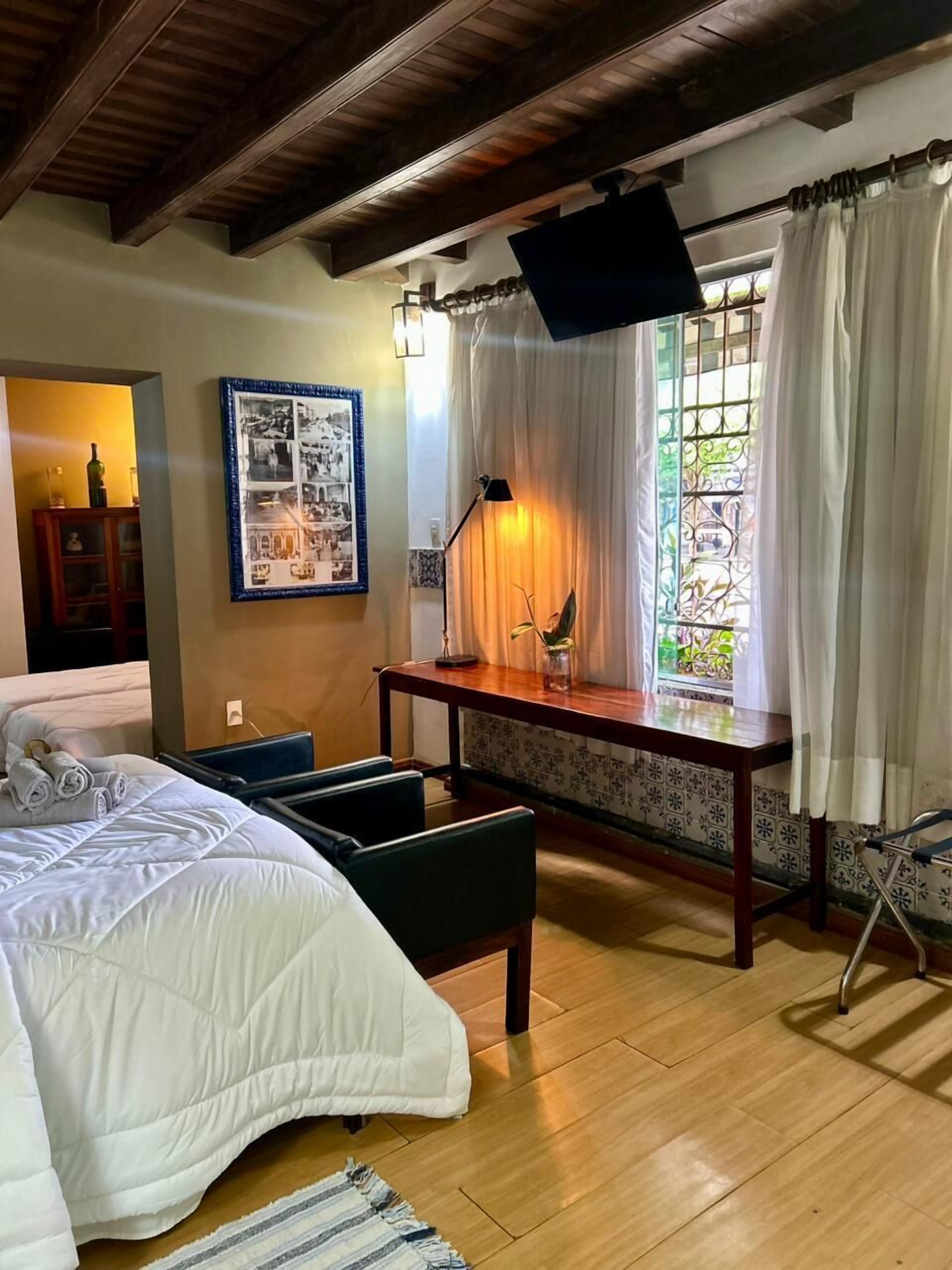 Habitación de hotel con cama, escritorio, silla, lámpara, ventana con cortinas e imagen enmarcada en la pared.